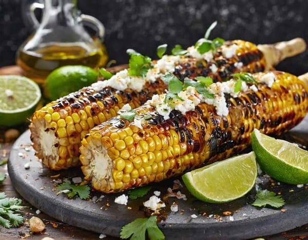 Charred grilled corn on the cob, kernels glistening with olive oil, topped with lime wedges and crumbled feta cheese.