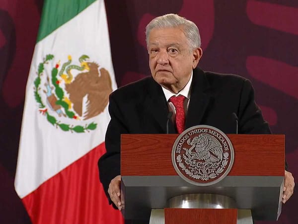 AMLO stands at a podium with the Mexican flag behind him, speaking into a microphone.