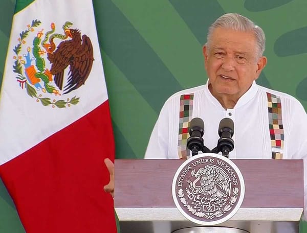 AMLO stands at a podium during his morning press conference, with microphones and the Mexican flag visible.