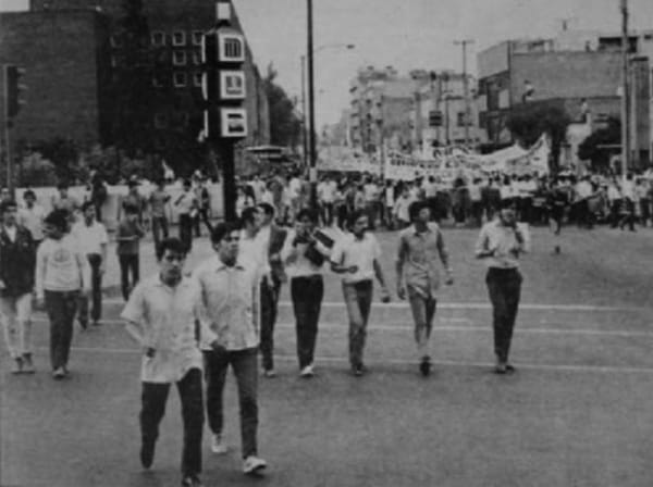 Black and white photo of a large crowd of students marching on a wide street, some carrying protest signs.