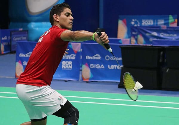 A badminton player in a red jersey swings their racket to hit a shuttlecock on a brightly lit court.